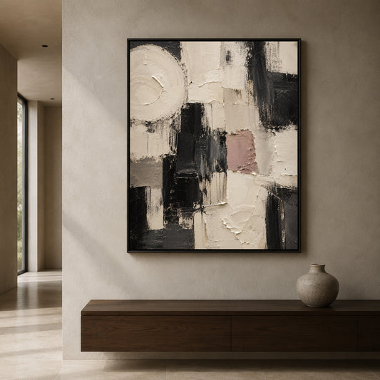 Abstract framed wall art with black, white, gray, and muted blush brushstrokes, displayed on a beige wall above a dark wood console with a ceramic vase.
