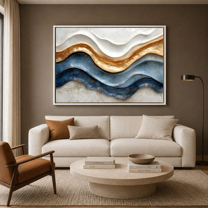 A large framed abstract wall art piece with flowing wavy bands of cream, gold, and deep blue hangs above a light beige sofa in a modern living room. The room also includes a brown accent chair, a round stone coffee table, books, and a floor lamp.