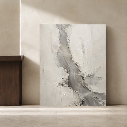 An abstract canvas painting with textured gray and off-white brushstrokes, leaning against a beige plaster wall in a minimalist interior setting.