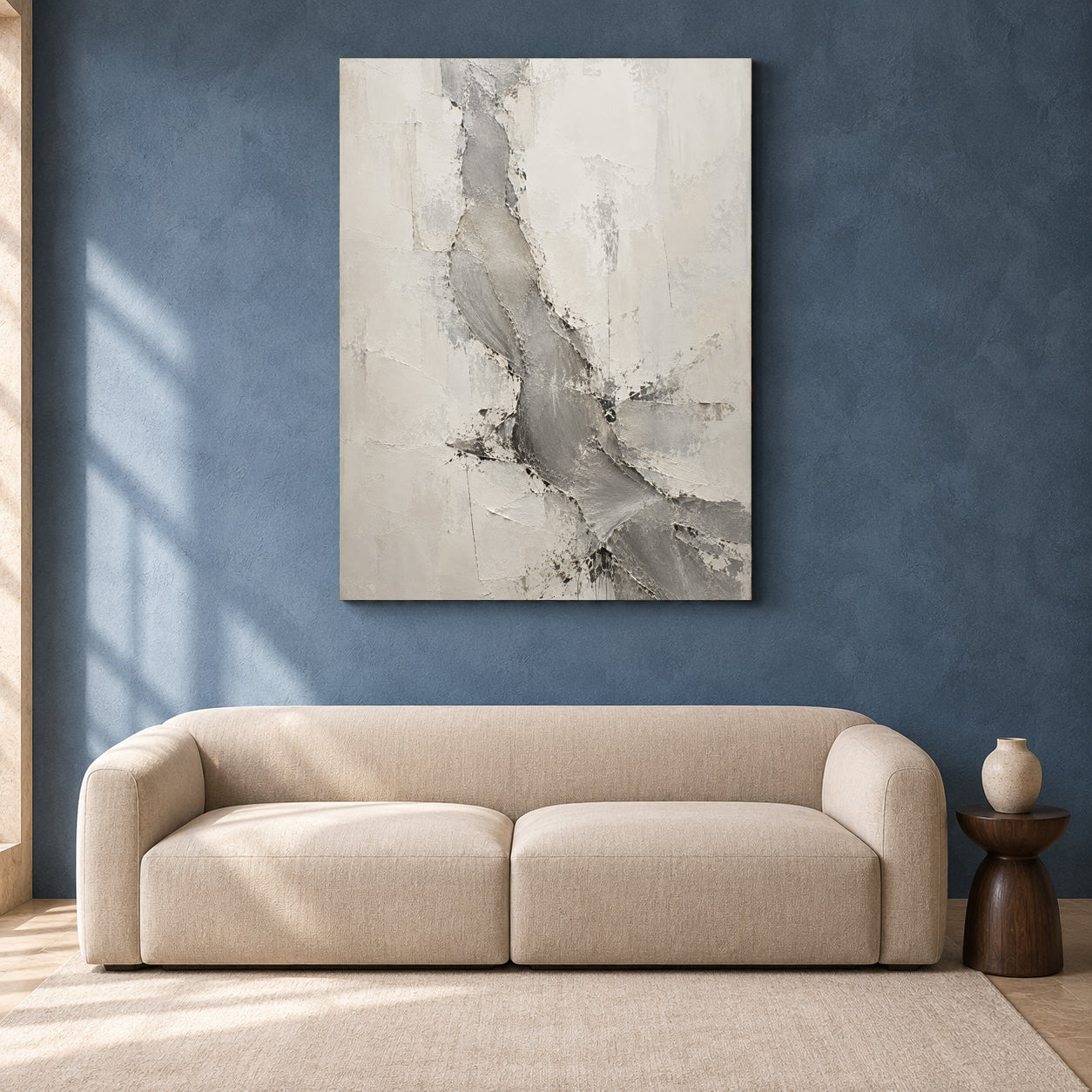 An abstract canvas wall art print with textured gray and off-white brushstrokes, displayed on a textured blue wall above a modern beige sofa in a styled living room setting.