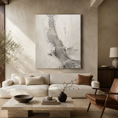 A large square abstract canvas print with gray and white textured brushstrokes hangs on a beige plaster wall above a cream-colored sectional sofa in a modern, sunlit living room.