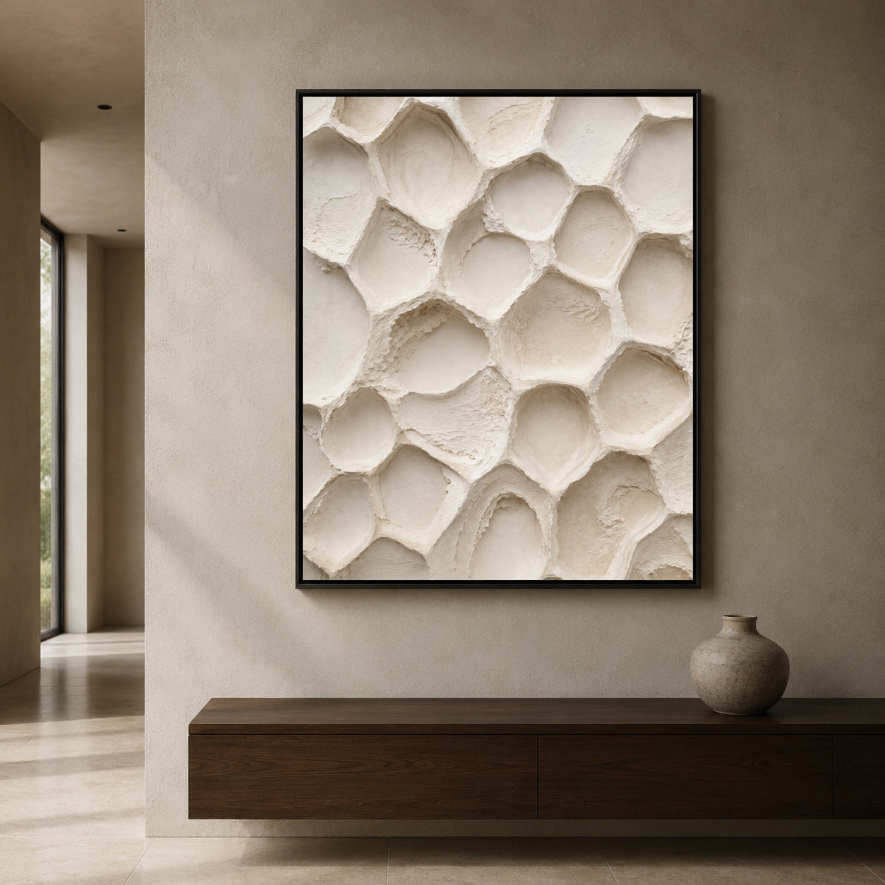A framed abstract wall art print featuring a textured, organic honeycomb-like pattern in warm off-white and cream tones, displayed on a beige wall above a dark wood console table with a small vase.