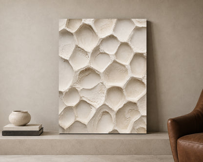 A large vertical canvas print featuring a 3D textured honeycomb or ionic lattice pattern in warm off-white and cream tones, displayed leaning against a neutral beige wall on a concrete ledge next to a brown leather armchair and a small ceramic vase.
