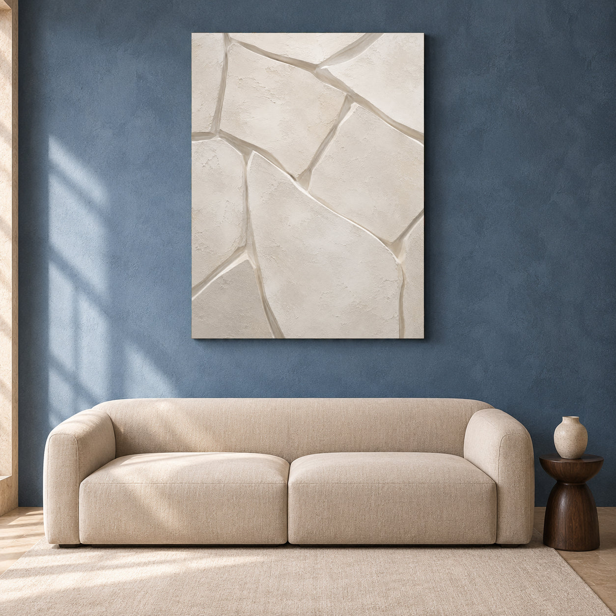 A large vertical canvas wall art print with an abstract cracked stone pattern in warm off-white and beige tones, displayed on a textured blue wall above a modern beige sofa in a styled living room setting.