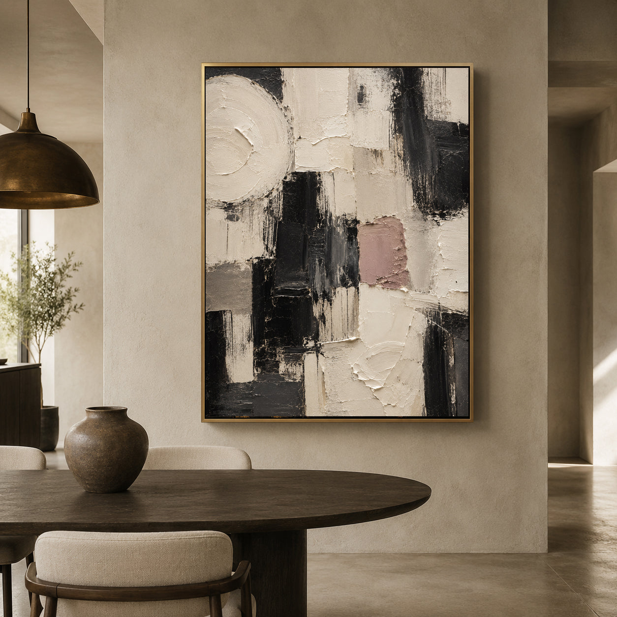 An abstract canvas painting with thick black, white, and gray brushstrokes and a muted dusty pink block, framed in gold and hanging on a beige wall above a dark wood dining table in a modern interior.