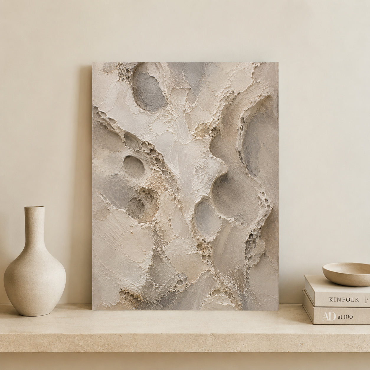 Abstract canvas wall art with textured organic shapes in warm beige, taupe, and gray tones, displayed leaning against a cream wall on a stone shelf beside a matte vase and stacked books.