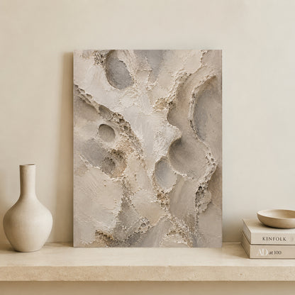 Abstract canvas wall art with textured organic shapes in warm beige, taupe, and gray tones, displayed leaning against a cream wall on a stone shelf beside a matte vase and stacked books.