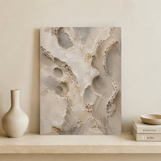 Abstract canvas wall art with textured organic shapes in warm beige, taupe, and gray tones, displayed leaning against a cream wall on a stone shelf beside a matte vase and stacked books.