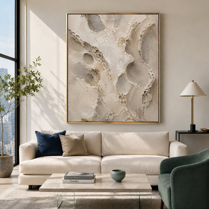 A large framed abstract canvas print with textured beige, cream, and gray organic shapes hangs on a light beige wall above a cream sofa in a modern living room setting.