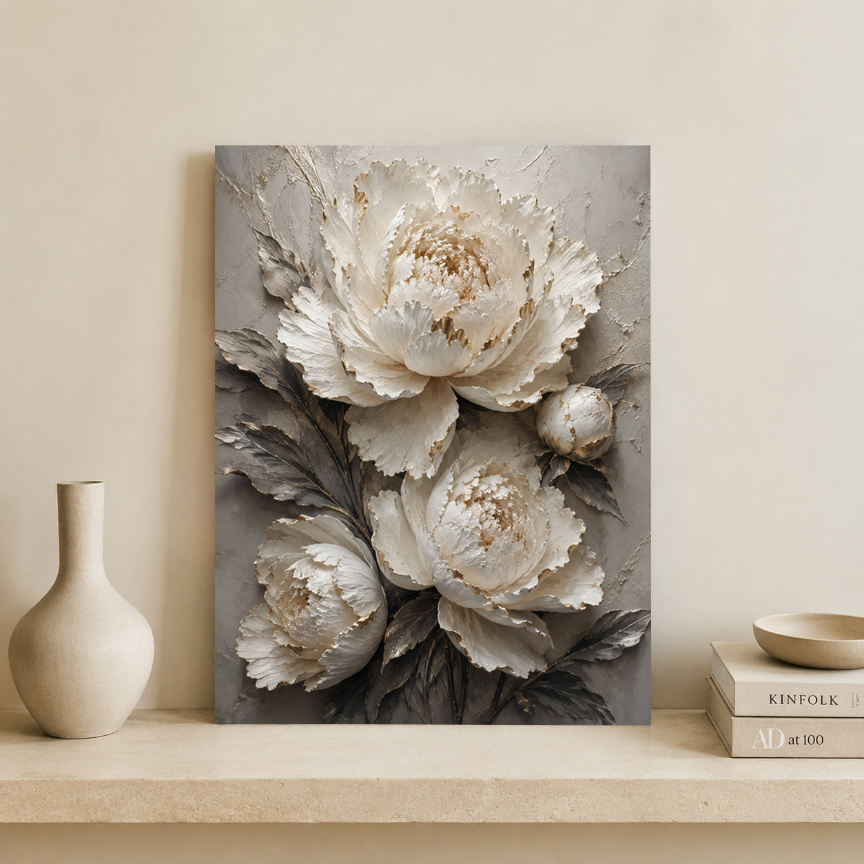 A vertical canvas wall art print featuring large textured white and gray peony flowers with gold-toned highlights, displayed leaning against a beige wall on a shelf beside a ceramic vase and books.