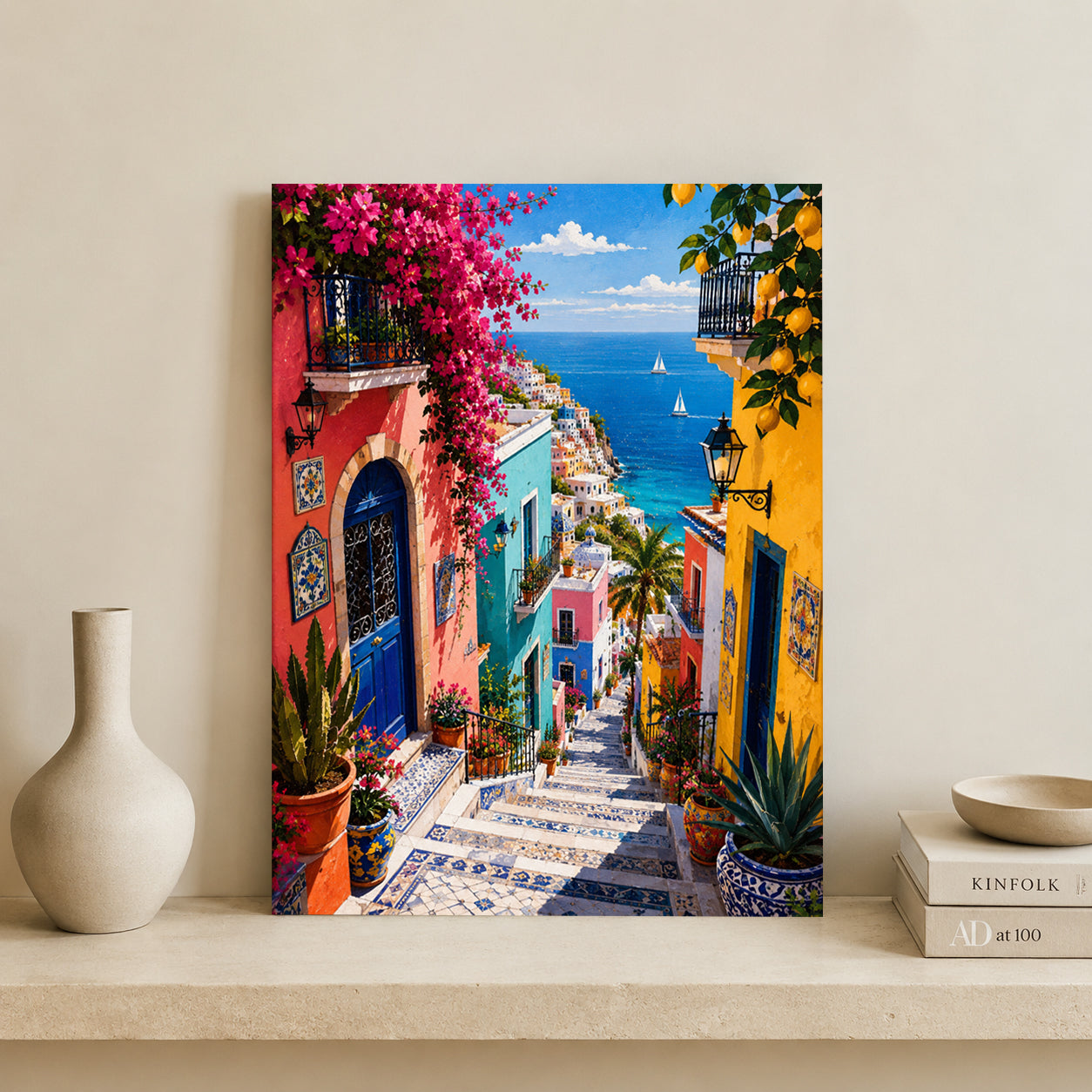 A vertical canvas print depicting a vibrant Mediterranean coastal street with colorful buildings, pink bougainvillea, tiled steps, and a blue sea, displayed on a shelf next to a white vase and books against a beige wall.