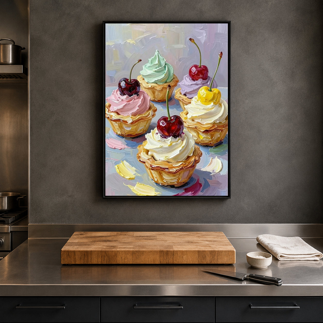 A framed canvas art print depicting a colorful still life of five iced tartlets with cherries, hanging on a dark gray kitchen wall above a wooden cutting board and stainless steel counter.