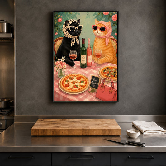 A framed wall art print hanging on a dark gray kitchen wall above a stainless steel counter. The artwork depicts two stylish cats seated at a table with wine glasses, bottles, pizza, a pink handbag, and flowers.