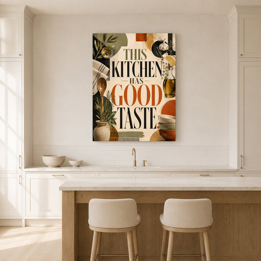 A vertical canvas wall art print featuring the text 'THIS KITCHEN HAS GOOD TASTE' in black and orange lettering, surrounded by retro kitchen illustrations, hanging on a white kitchen wall above a wooden island with two stools.