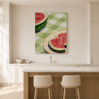 A vertical canvas wall art print depicting watermelon slices on a green and white checkered tablecloth, displayed in a bright, modern kitchen above a wooden island and two bar stools.