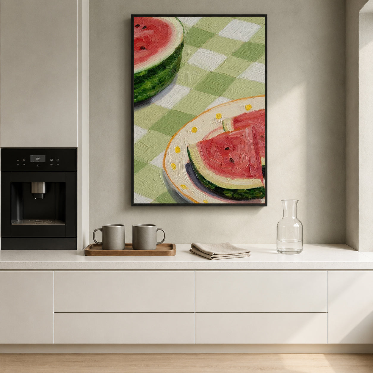 A framed vertical art print depicting watermelon slices on a green-and-white checkered tablecloth, hanging on a light gray wall above a white kitchen counter with mugs, a glass bottle, and a coffee machine.